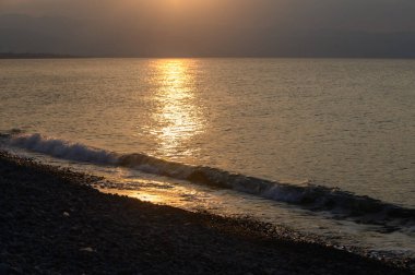 Altın güneş ışığı, kayalık bir plaja yuvarlanan yumuşak dalgalarda parlıyor ve alacakaranlık yaklaşırken huzurlu bir ortam yaratıyor. Ufuk, akşam gökyüzünde güzel renkleri harmanlıyor..