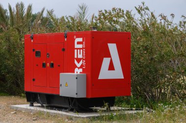 Gaziveren Kıbrıs Rum Kesimi -17.03.2025 Canlı bir kırmızı jeneratör yemyeşil bitkilerin arasına sıkışmış beton bir tabana yerleştiriliyor. Gri bulutların altında sakin bir açık hava ortamında güç sağlamaya hazır..