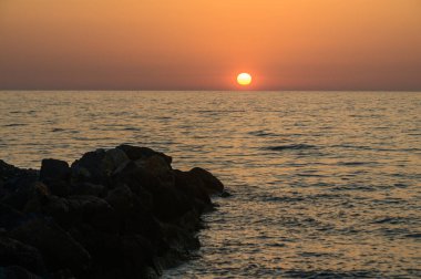 Kıbrıs 'taki Akdeniz kıyılarında nefes kesici bir günbatımı. Gökyüzü turuncu, kırmızı ve mor renklerle dolu. Güneş batarken sakin sular ve sahil şeridi üzerine büyülü bir parıltı saçıyor..