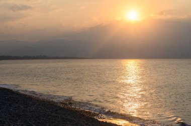 Güneş ufkun altına batarken, yumuşak altın ışınlar sakin sulardan yansıyarak çakıl taşlı bir sahili aydınlatıyor. Hafif bir esinti alacakaranlıkta huzurlu atmosferi karıştırır..