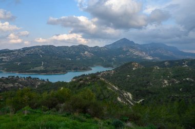 Parlak bulutlar yeşil tepeler ve görkemli dağlarla çevrili sakin bir gölün üzerinde süzülür yumuşak bir öğleden sonra ışığı altında, doğada barışçıl bir atmosfer yaratır..