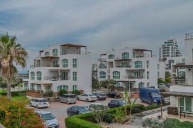 Bereketli yeşilliklerle çevrili bulutlu bir gökyüzüne kurulmuş zarif beyaz binalar ve huzurlu bir yerde park edilmiş araçlar..