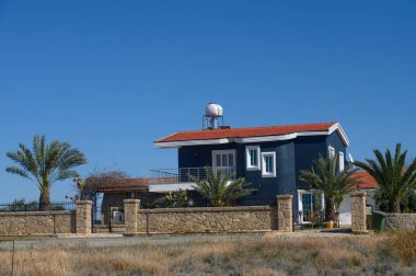 Güzel mavi bir ev gururla duruyor. Etrafı yemyeşil palmiye ağaçları ile çevrili. Parlak kırmızı bir çatı öğleden sonra güneşini yansıtıyor. Huzurlu kırsal bölgeler cazibesini arttırarak sakin bir atmosfer yaratıyor..