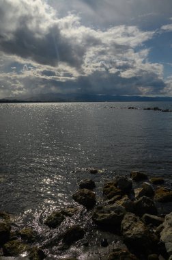 Sakin sular güneş ışığının parıltısını yansıtırken bulutlar yukarıda toplanıyor. Kıyı boyunca uzanan kayalar kara ve deniz arasında doğal bir sınır oluşturuyor. Yansımayı davet ediyor..