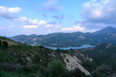 Yuvarlanan tepeler, alacakaranlığın yumuşak tonlarını yansıtan sakin bir gölü beşiyorlar. Kabarık bulutlar havada süzülür, yemyeşil manzaraya gölgeler bırakır. Atmosfer huzurlu, davetkar bir keşif..