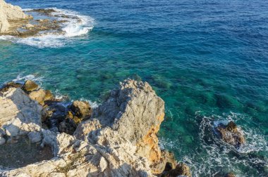 Dalgalar engebeli kayalara hafifçe çarparken, canlı mavi su güneş ışığı altında parlarken, sükunet yaratır..
