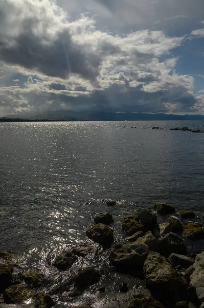 Sakin sular güneş ışığının parıltısını yansıtırken bulutlar yukarıda toplanıyor. Kıyı boyunca uzanan kayalar kara ve deniz arasında doğal bir sınır oluşturuyor. Yansımayı davet ediyor..