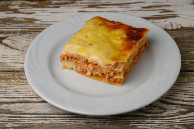 İçinde bol et sosu ve erimiş peynir bulunan tuzlu lazanya katmanları, temiz beyaz bir tabakta dinleniyor. Kırsal ahşap masa ev yapımı yemeğe sıcaklık katar ve açlığı davet eder..