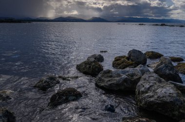 Sakin gölün üzerinde kara bulutlar beliriyor. Güneş batmaya başladığında kayalık kıyı şeridine gölgeler süzülüyor..
