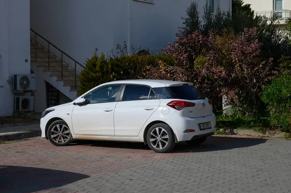 Gaziveren Kıbrıs Rum Kesimi - 01.03.2025 Parlak beyaz bir hatchback, yemyeşil bitkilerle çevrili çağdaş bir yapının arka planına sessizce dayanır.
