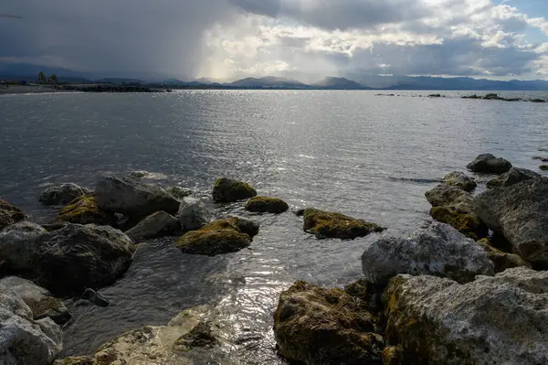 Dalgalar kıyı boyunca uzanan engebeli kayalara hafifçe çarpar, bulutları aşan güneş ışığını yansıtır. Dağlar görkemli bir şekilde yükselir, dingin güzelliği arttırır..