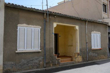 Ev, yıpranmış duvarlar ve klasik panjurlarla çerçevelenmiş güneşli bir girişe sahip, sakin bir caddede sakin bir sükunet hissi somutlaştırıyor..