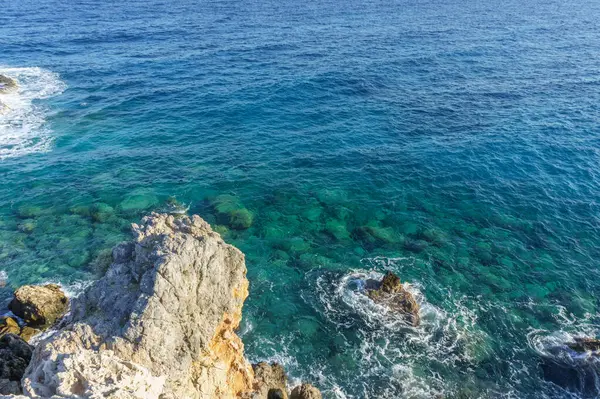 Kristal berrak sular güneşin altında parıldıyor, sakin sahil boyunca engebeli kayaları kucaklıyor..