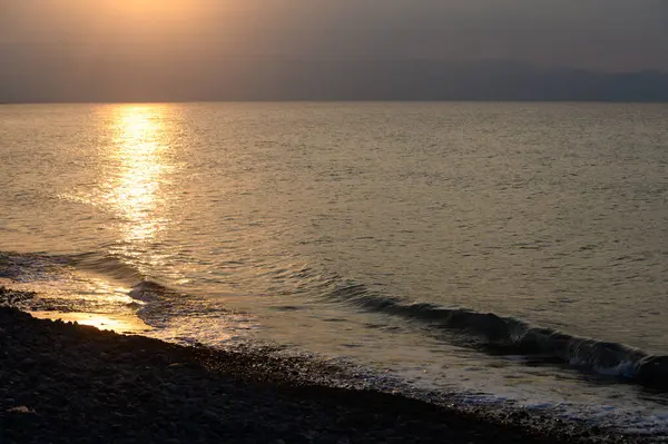 Güneş ufkun altına batarken suyun yüzeyinde altın renkler dans ediyor ve sakin bir ortam yaratıyor. Yumuşak dalgalar çakıl taşlarına çarpar, rahatlatıcı bir melodi yaratır..