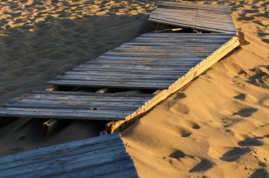 Yıpranmış ahşap bir patika sessizce durur, yumuşak kumda yarı batmış, sıcak bir günbatımı parıltısıyla aydınlanmış..