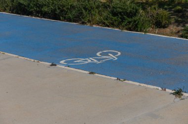 Canlı mavi bir bisiklet yolu, beyaz bir bisiklet sembolü ile işaretlenmiş huzurlu bir park boyunca uzanır. Güneş ışığı, açık hava maceraları için mükemmel olan yeşil yol boyunca yumuşak gölgeler bırakır..