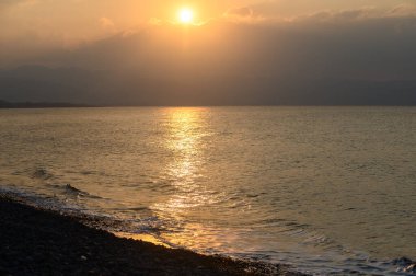 Güneş ufukta batarken dalgalar hafifçe çakıllı bir kıyıya çarpar ve su üzerinde altın ve turuncu renklerin nefes kesici bir yansımasını yaratır. Doğanın güzelliği bu huzurlu anda ortaya çıkar..