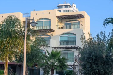 Çağdaş binalar bereketli palmiye ağaçları ile bezenmiş sıcak güneş ışığında güneşlenen, sakin bir sahil atmosferini tasvir eden..