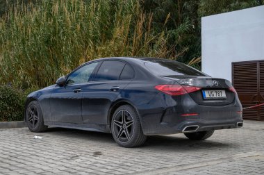 Gaziveren Kıbrıs Rum Kesimi -28.02.2025 Pürüzsüz, siyah, lüks bir sedan, bulutlu gökyüzünün altındaki yemyeşil bitkilerle çevrili, asfaltlı bir alanda sessizce duruyor..