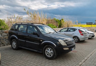 Gaziveren Kıbrıs Rum Kesimi - 28.02.2025 Siyah bir SUV, bulutlu bir günde koyu bulutlar ve canlı sarı alanlar tarafından çerçevelenmiş küçük arabaların arasına park edilmiştir..
