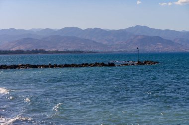 Bereketli mavi sular, sakin bir gökyüzü ile harmanlanır. Sıcak bir güneşin altında ufka doğru solan dağlar sergilenir..