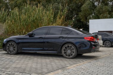 Gaziveren Kıbrıs Rum Kesimi -28.02.2025 Şık, siyah bir sedan, canlı yeşillik ve çağdaş mimariyle çevrili kaldırımda sessizce dinleniyor.