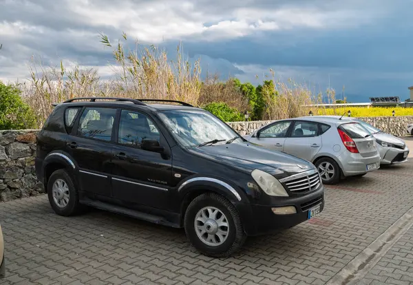 Gaziveren Kıbrıs Rum Kesimi - 28.02.2025 Siyah bir SUV, bulutlu bir günde koyu bulutlar ve canlı sarı alanlar tarafından çerçevelenmiş küçük arabaların arasına park edilmiştir..