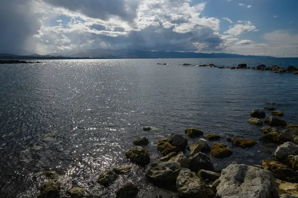 Dalgalar kayalar ve yosunlarla dağılmış engebeli bir kıyı şeridine nazikçe çarpar. Bulutlarla dolu dramatik bir gökyüzü değişen gölgeler oluşturur, günbatımında sakin bir atmosfer yaratır..