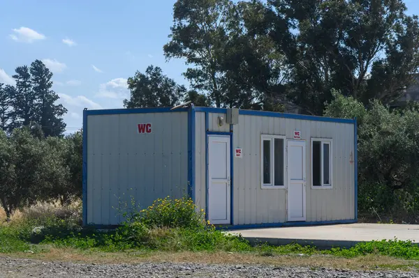 Uzun ağaçlar ve canlı bitkilerin arasında korunan bir konteynır tuvaleti tek başına duruyor. Güneş ışığı yaprakların arasından süzülerek bu huzurlu yerde barışçıl bir atmosfer yaratır..