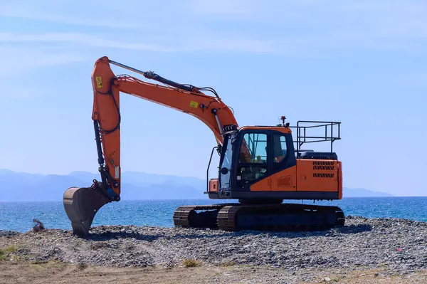 Akdeniz 'in mavi suları ve engebeli kıyı şeridi tarafından çerçevelenen Kıbrıs' taki bir kıyı şeridinde turuncu bir kazıcı boşta duruyor