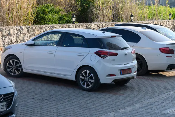 Gaziveren Kıbrıs Rum Kesimi 03.02.2025 Taş duvarlar ve yeşillik yakınlarındaki parlak güneş ışığı altında gümüş bir sedanın yanına park etmiş parlak beyaz bir hatchback..
