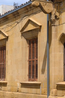 Güneş ışığı, karmaşık mimari detaylar, ahşap kepenkler ve klasik bir taş cephe içeren güzel tarihi bir binaya gölge düşürüyor. Canlı şehir atmosferi ortama cazibe katıyor..