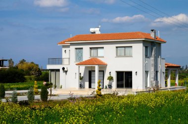 Ilık bir günde, berrak mavi bir gökyüzünün altında yemyeşil ve canlı çiçeklere yuva kurmuş parlak, ferah bir ev..