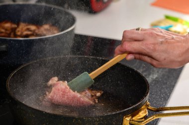 Bir el, iyi donanımlı bir mutfakta, sıcak bir tavada cızırdayan eti ustalıkla çevirir, aromatik buhar saçar..