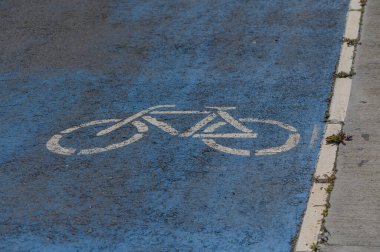Canlı bir mavi bisiklet yolu işareti güneşli bir şehir caddesinde göze çarpıyor. Bu açık gösterge, hareketli şehirde bisiklet sürmeyi yeşil bir ulaşım tercihi olarak teşvik eder.