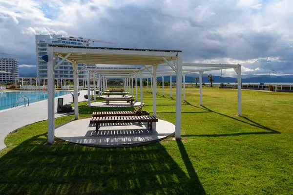 Gölgeli ahşap banklar, yemyeşil çimenler ve canlı bulutlar tarafından çerçevelenmiş dingin bir havuz alanı hizasına gelir..