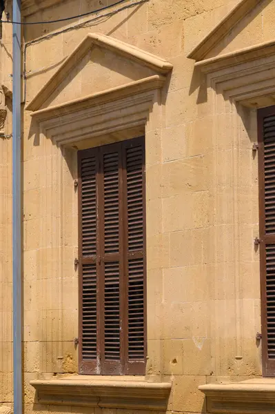 Sıcak güneş ışığının tadını çıkaran antik bir taş yapı kaba dokulu duvarları ve koyu ahşap kepenkleri olan köy cazibesini sergiliyor. Renkler sakin bir atmosfer yaratıyor..