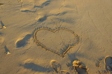 Bir kalp çizgisi, kumsalda güneş doğarken ayak izleriyle çevrili yumuşak kumda duruyor..