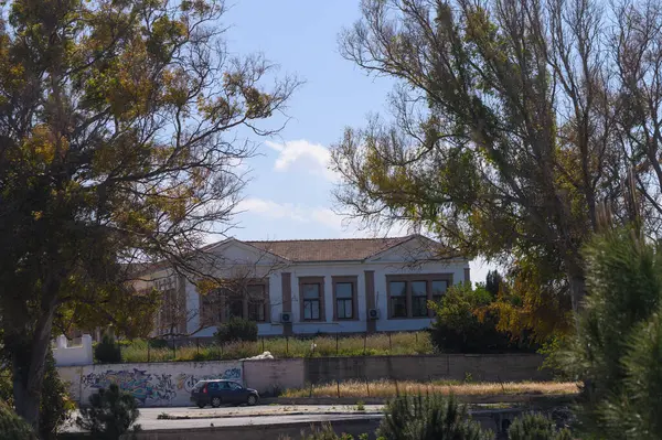 Eski, terk edilmiş bir bina uzun ağaçların arasında sessiz duruyor, solgun boya tarihini açığa vuruyor. Yukarıdaki mavi gökyüzü yalnızlık duygusunu artırıyor. Zamanla unutulan bir şehir ortamını yakalıyor..