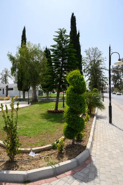 Parlak yeşillik sakin bir parkı dolduruyor, güneşli bir öğleden sonra açık mavi gökyüzü altında düzgün budanmış çalılar ve uzun ağaçlar sergiliyor. Aileler patikalarda rahatça yürümekten hoşlanırlar..