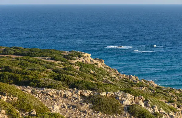 Engebeli kayalıklara bakan güneş sakin denizin üzerinde batarken yumuşak dalgalar kıyıya vurur..