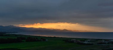 Güneş ufkun altına batarken, altın ışınlar engebeli kıyı dağlarını ve sakin denizi aydınlatıyor. Üzerimizde kara bulutlar sürükleniyor, akşam gökyüzünde dramatik bir kontrastlık yaratıyor..