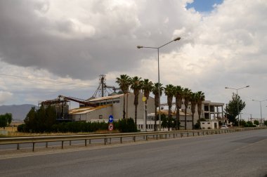 Kıbrıs 'ın kırsal kesiminde ağaçlar ve uzak dağlarla çevrili endüstriyel bir tahıl asansörü. Arkaplanda açık gökyüzü.