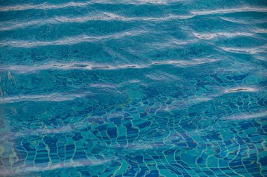 Yüzme havuzundaki berrak mavi su yüzeyinde güneş ışığı yansımaları var..