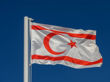 Kuzey Kıbrıs bayrağı hafif bulutlu mavi gökyüzüne karşı rüzgarda dalgalanıyor.
