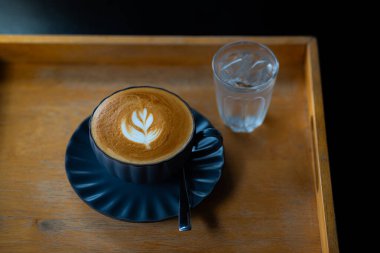 Sütlü kahvenin en üst görüntüsü, siyah bir fincanda latte sanatıyla, ahşap masada bir bardak buzlu suyla.