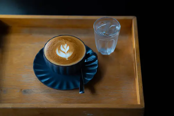 Sütlü kahvenin en üst görüntüsü, siyah bir fincanda latte sanatıyla, ahşap masada bir bardak buzlu suyla.