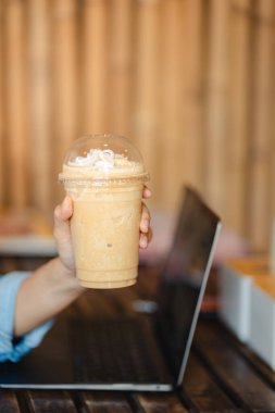 Ofisteki masada dizüstü bilgisayarlı buzlu kahve frappe bardağını kapat. Çalışma alanı kavramı..