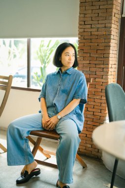 Şen şakrak esmer, kıvırcık pantolonlu ve gömlekli kadın gülümsüyor ve manzaraya bakıyor. Kot pantolonlu güzel bayan kafede sandalyede oturuyor.