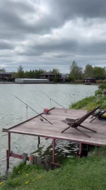 Arka planda bir göl ve olta kamışlarıyla ahşap bir balıkçı iskelesinin statik görüntüsü. Yüksek kaliteli FullHD görüntüler